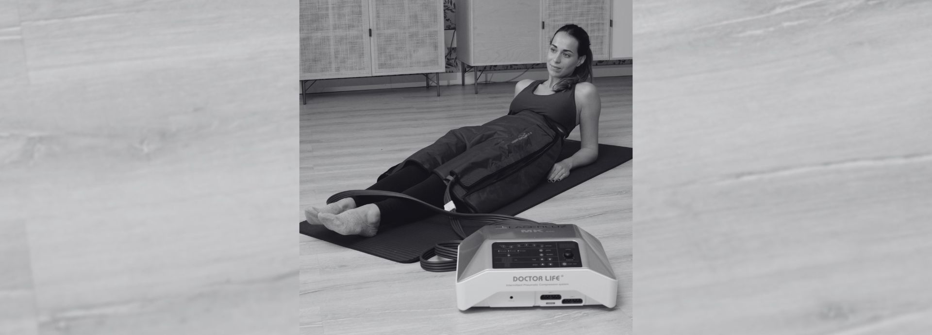 Foto en blanco y negro de una persona que recibe un tratamiento estético con el equipo PRESOTERAPIA ELIXIA LITE, tumbada en una esterilla de yoga sobre un suelo de madera.